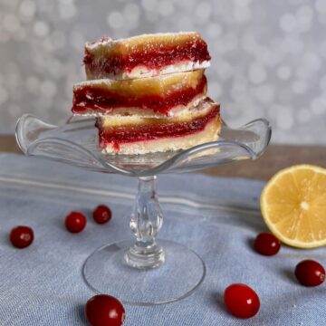 Cranberry Lemon Bars stacked on a glass cake stand with fresh cranberries and sliced lemon below on a blue napkin with white stripes.