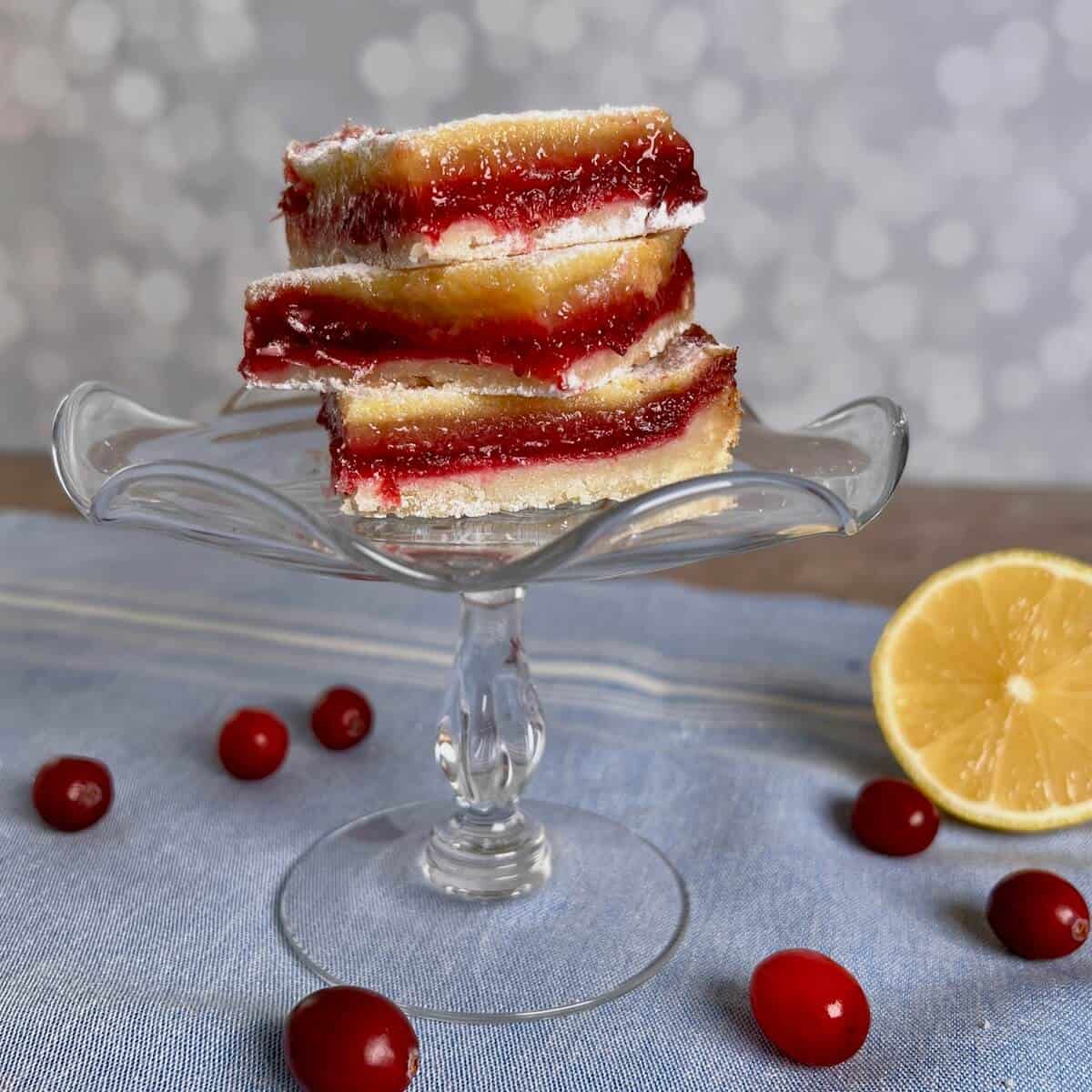 Cranberry Lemon Bars stacked on a glass cake stand with fresh cranberries and sliced lemon below on 